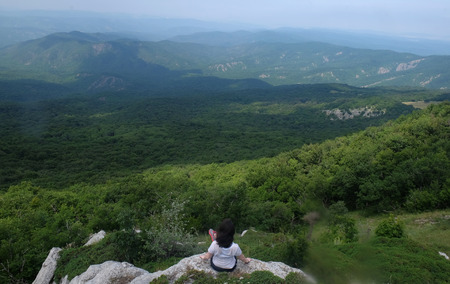 A young girl sits on edge cliff and looking at mountain landscapeの写真素材