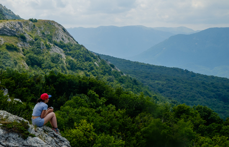 A young girl sits on edge cliff and looking at mountain landscapeの写真素材