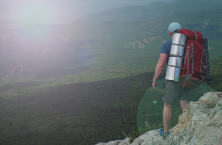Young man tourist with a big backpack standing on the edge of a precipice and looking at mountain landscapeの写真素材