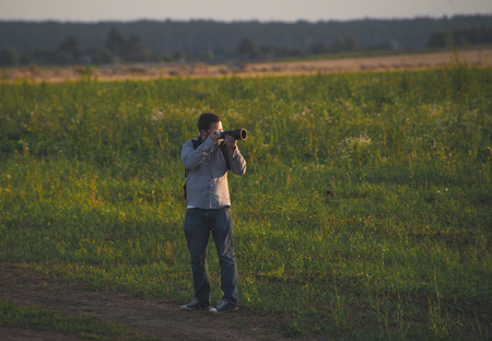 A young white man with a camera walking on the field and taking pictures.