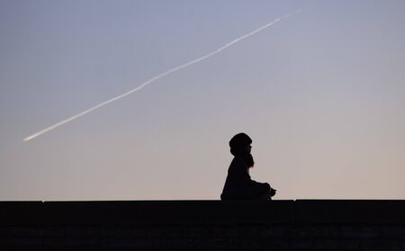 The silhouette of a meditating girl against the background of the condensation trail of a passing...