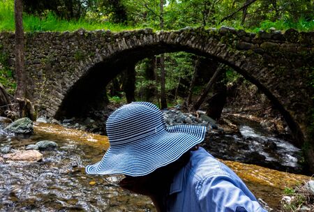 A girl in a wide-brimmed hat drinks water from a stream next to a medieval Venetian bridge in the center of the island of Cyprus.の写真素材