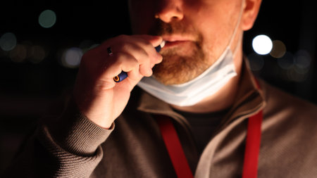 A white unshaven man with a medical mask lowered on his chin smokes an electronic cigarette