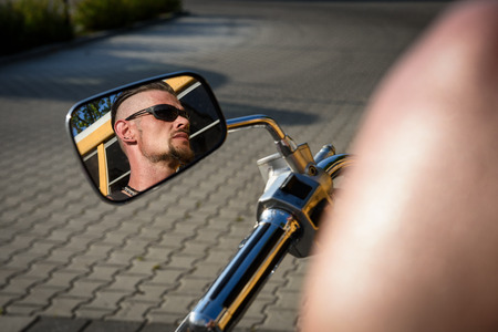 Tough guy with undercut, sparrow beard and sun glasses reflecting in the rear view mirror of his bikeの写真素材
