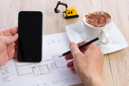Close-up of architect hands working on a blueprint and holding a smartphone. Cup of coffee and miniature extractor in the background. Blueprint was created by photographer.の写真素材