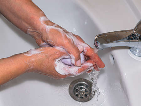 Women's hands in the process of washing with soap and water pouring from a chrome-plated tap into a white sink.の写真素材