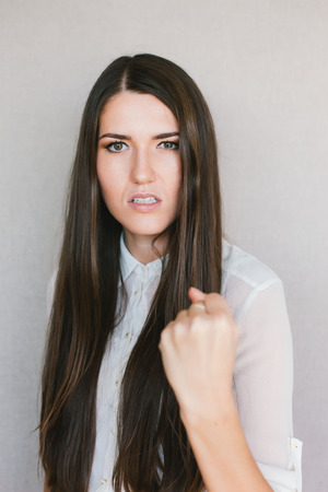 Half-length portrait of beautiful dark-haired young angry woman wearing white blouse   showing us her fistsの写真素材