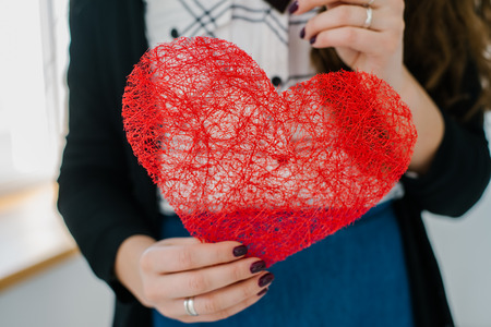 happiness, health, people, holidays and love concept - smiling young woman in white t-shirt holding red heart against the windowの写真素材
