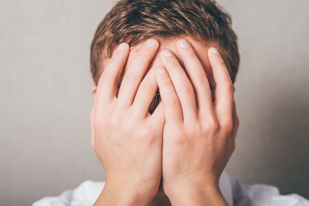 Shocked and terrified. Portrait of young man covering his face by hand and looking at camera while standing against grey backgroundの写真素材
