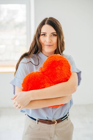 Portrait of a  beautiful young brunette hugging red heart shaped pillow  against the window.の写真素材