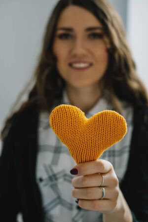 portrait of attractive  caucasian smiling woman with long curly hear brunette with yellow heart in hands near the windowの写真素材