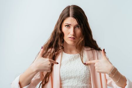 long-haired beautiful young brunette woman with surprise points to itself fingers, isolated on a white backgroundの写真素材