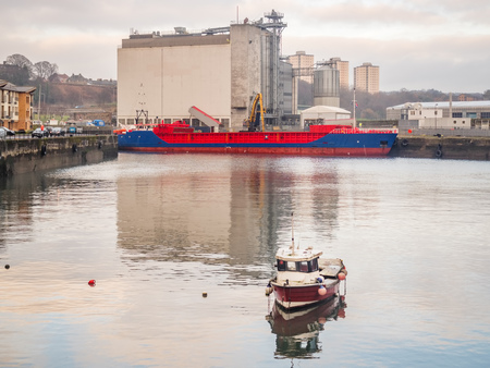 Blue and red Dutch small ship under grain discharging operation in harbor of Kirkcaldy with a small boat in frontの写真素材