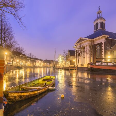 Old Dutch city Schiedam evening landscape during freezing weather with reflections in a canal, old barges and windmill and ice on waterの写真素材