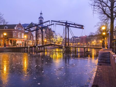 Old Dutch city Schiedam evening landscape during freezing weather with reflections in a canal, old bridge and ice on waterの写真素材