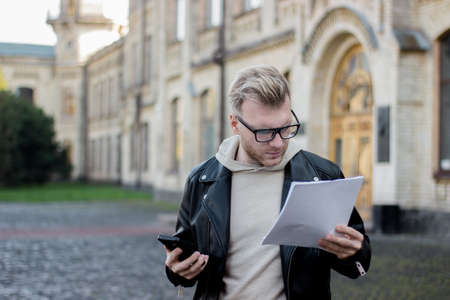 A smart guy in casual clothes holds phone and looks at papers while standing next to the university....
