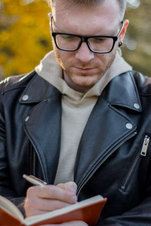 Smart guy writer in casual clothes makes notes in a notebook, a successful student in black glasses and a jacket does homeworkの写真素材