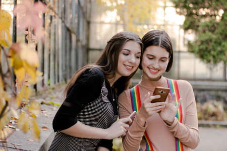 Two beautiful studentslooking at the phone in the autumn park. Girls communicate online via video...
