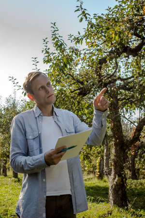 Guy in casual clothes is holding a pen with papers and looking right against a background of trees....