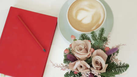 Cappuccino in a cup with a heart on the crema. Coffee with a red notebook and a small bouquet on the table. View from aboveの写真素材