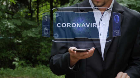 Unrecognizable businessman activates conceptual HUD holograms on smartphone with text Coronavirus. Bearded man in a white shirt and a jacket with a holographic screen on a background of green treesの写真素材