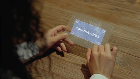 Unrecognizable woman working on a futuristic tablet with a hologram text Coronavirus. Womens hands with future holographic technology at a wooden tableの写真素材