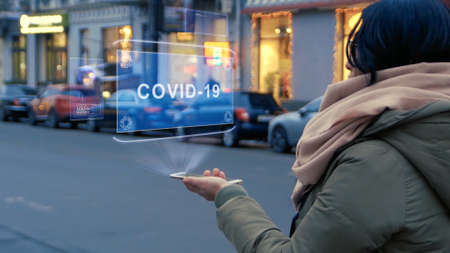 Unrecognizable woman standing on the street interacts HUD hologram with text COVID-19. Girl in warm clothes with a scarf uses technology of the future mobile screen on background of night cityの写真素材