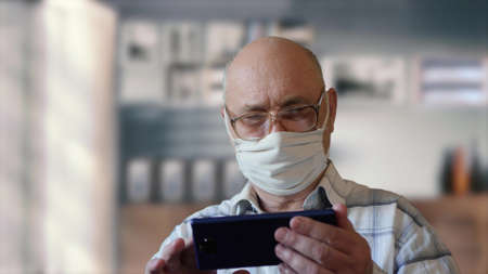 Elderly man in isolation at home wearing a handmade virus protection mask uses phone. Reading news...