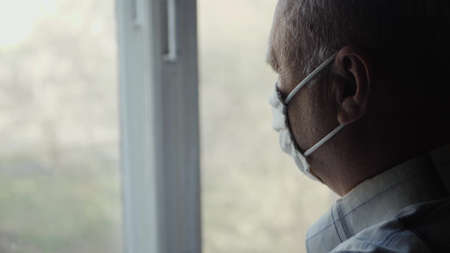 Old man in a protective mask on his face looks out the window copy space. Quarantine Restriction Concept. Elderly man indoors during an epidemicの写真素材
