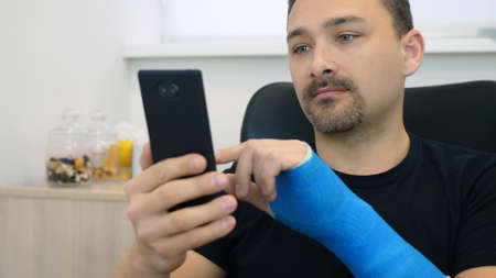 Handsome middle-aged man in black t-shirt with broken wrist surfs the internet at home. Male hand,...