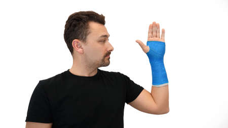 Handsome middle-aged bearded man in a black T-shirt looks at his hand wrapped in a plaster cast on a...