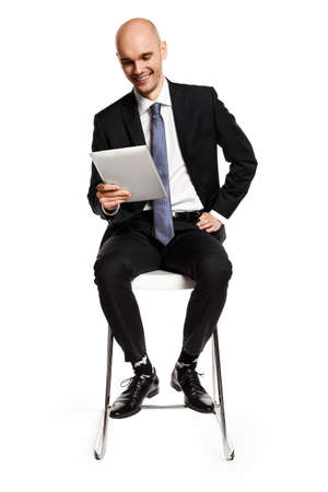Cheerful young businessman sitting on a chair and looking at digital tablet. Isolated on white background.の写真素材