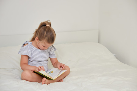 Cute little girl sitting on bed at home and reading book with fairy talesの写真素材