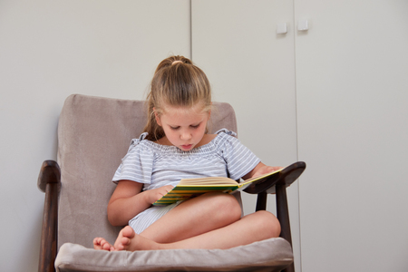 Cute little girl sitting in cushioned armchair at home and reading book with fairy talesの写真素材