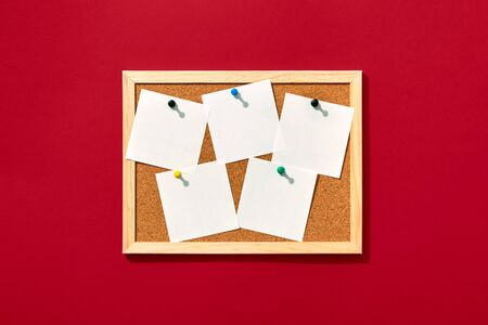 Cork board with wooden frame with five pinned blank notes on red background, view from above with...