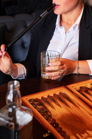 Unrecognizable young woman in white blouse and suit drinking whiskey, smoking hookah and playing...