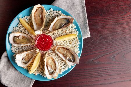 Platter with half-dozen of fresh opened oysters with sauce and slices of lemon on white stones on wooden table. Top view with place for text.の写真素材