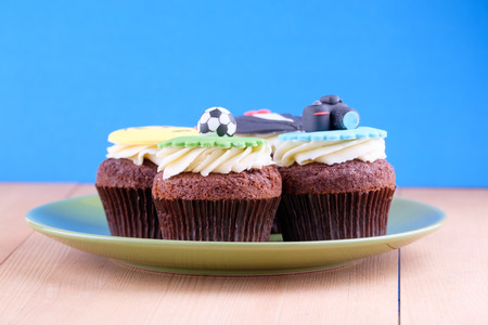 Delicious cupcakes with icons of ball, tuxedo, smiley and camera on it with green plate on wooden desk and blue backgroundの写真素材