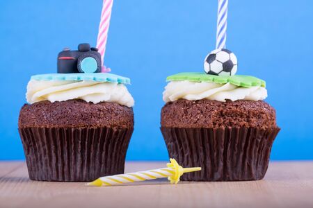 Delicious birthday cupcakes with icons of ball and camera on it and candles on wooden desk and blue backgroundの写真素材