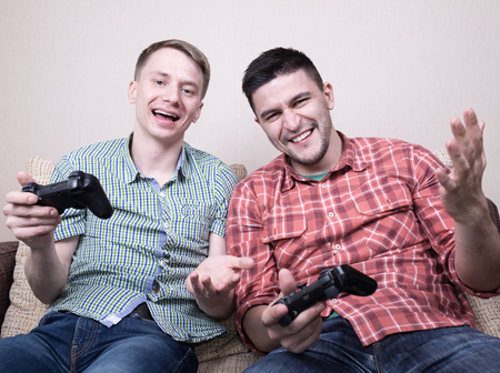 Two young guys playing video games at home, holding controllers and sitting on sofaの写真素材