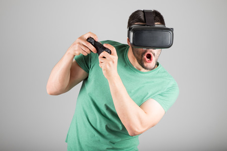 Young man playing with virtual reality glasses and video game controller isolated on a gray...