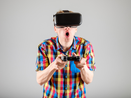 Young man playing with virtual reality glasses and video game controller isolated on a gray...