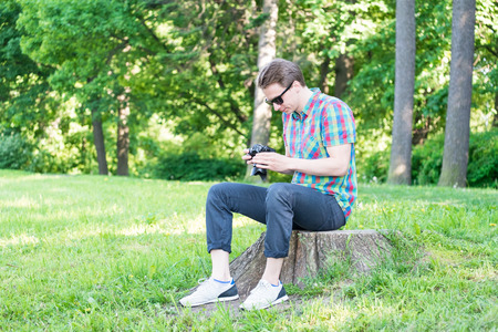 Young handsome guy in colorful shirt looking  on pictures in camera and sitting on stump in park