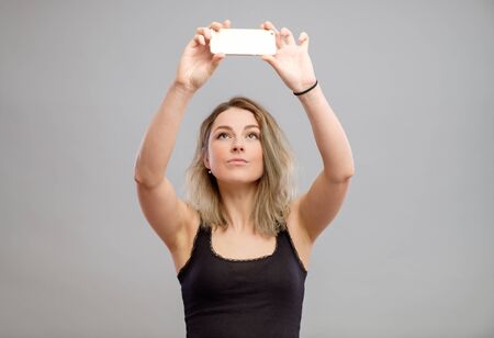 Expressive young woman taking pictures through cellphone, over gray backgroundの写真素材