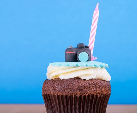 Delicious birthday cupcake with realistic camera icon on it and candle on wooden desk and blue backgroundの写真素材