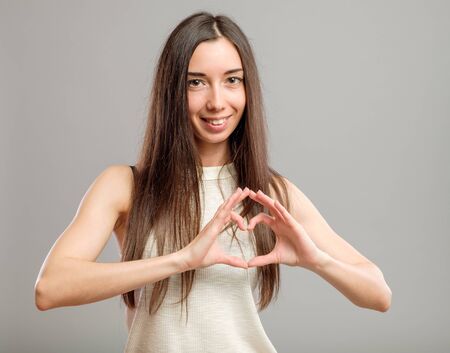 Beautiful young woman over a gray background showing a heart shape with her handsの写真素材