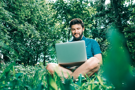 Happy cheerful hipster man with a laptop sitting outdoors on green grass. Freedom  concept