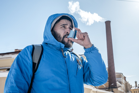 Portrait of young man using asthma inhaler outdoor with factory chimney on backgroungの写真素材