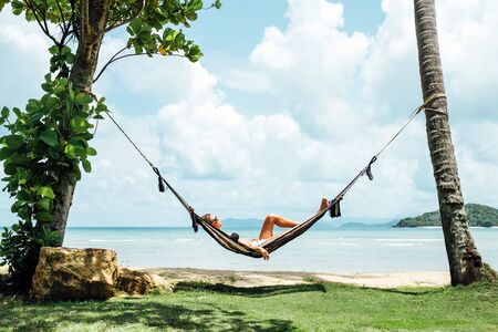 Summer vacations concept. Happy woman in black bikini relaxing in hammock on tropical beachの写真素材
