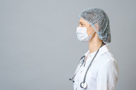 Portrait of young female doctor in protective medical mask on her face and cap on her head. Isolated on gray backgroundの写真素材